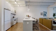 Located at 5050 N Broadway St in Chicago's vibrant Uptown neighborhood, this single-floor loft-style unit blends industrial character with modern design. Polished concrete floors and exposed structural beams run throughout the space, creating a cohesive, urban aesthetic that sets the tone from entry to living area.

The open-concept kitchen is anchored by a central island with a white quartz countertop, an integrated sink, and bar seating for multiple guests. A stainless gas range, full-size refrigerator, white upper cabinetry, and open shelving complete the well-appointed cooking space. Overhead, a combination of track lighting and pendant fixtures provides targeted illumination across the kitchen and dining area.

The living room offers a comfortable and stylish retreat, with large windows allowing natural light to filter in. A wall-mounted mini-split system ensures year-round climate control, while the open layout accommodates generous seating arrangements with ease.

The bathroom showcases a refined, contemporary design featuring a sleek floating vanity in a muted blue-gray finish with a white quartz countertop, a mirrored medicine cabinet, and a glass-enclosed walk-in shower with full-height white subway tile and a rain showerhead. The polished concrete flooring carries seamlessly into this space, reinforcing the unit's unified design language throughout.