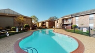 Westridge Gardens, located at 2501 Westridge St in Houston, Texas, is a well-established residential community characterized by its classic brick exterior architecture and thoughtfully maintained grounds. The community is spread across a single level and organized around a welcoming central outdoor area that serves as a gathering hub for residents.

At the heart of the community lies a generously sized freeform swimming pool with vivid turquoise water, enclosed by a black iron fence and surrounded by a brick-trimmed pool deck. Adjacent to the pool, a covered patio area offers comfortable outdoor seating with colorful tables and chairs shaded by large red market umbrellas, creating an inviting space for relaxation and socializing under the Houston sun.

The exterior of the community features warm brick facades, covered walkways, and balconies that contribute to the neighborhood's traditional residential character. Mature trees and manicured shrubs are distributed throughout the property, providing greenery and shade. Red mulched landscaping beds line the walkways and building entrances, adding a polished, well-kept appearance.

The on-site leasing office, marked by a "Welcome to Westridge Gardens" sign, provides a convenient point of contact for residents and visitors. Ample surface parking is accessible throughout the community, and wide driveways ensure easy navigation across the property.