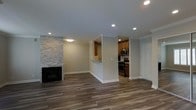 This single-floor space combines a spacious living area and a well-appointed kitchen in a cohesive, open layout. The living room serves as the focal point of the space, anchored by a striking floor-to-ceiling stacked stone fireplace that adds texture and visual interest to the neutral, gray-toned walls. Rich, dark hardwood-style flooring runs throughout, providing a warm contrast to the light color palette. Crown molding along the ceiling adds a refined architectural detail, while recessed lighting ensures consistent, even illumination throughout the space.

A half-wall partition separates the living area from the kitchen, creating a semi-open connection between the two spaces. The kitchen features warm-toned wood cabinetry, stainless steel appliances including a range and microwave, and a decorative tile backsplash that introduces subtle pattern and depth. A pass-through opening with counter space allows for casual interaction between the kitchen and the main living area.

The overall ambiance is clean and modern, with a neutral palette of grays and warm browns creating a versatile and inviting atmosphere throughout.