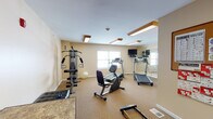 Located at 1320 Southwestern Blvd in Buffalo, NY, this single-floor fitness center offers a well-equipped workout space designed to support a variety of exercise routines. The room features warm beige walls, neutral carpeted flooring, and a bright, clean atmosphere illuminated by wood-trimmed ceiling light fixtures that lend a warm tone to the space.

The main workout area is outfitted with multiple cardio machines, including treadmills and a recumbent stationary bike, along with a multi-function cable strength training station and a rowing machine. A large wall-mounted mirror expands the sense of space and allows users to monitor their form during exercise. A wall-mounted flat-screen television is positioned for easy viewing from the cardio equipment, and a stability ball rounds out the functional fitness offerings.

Natural light filters in through windows fitted with horizontal blinds, providing a pleasant and energizing environment. A seating area along one side of the room includes a tufted black bench and a small table, offering a spot to rest or review workout notes. The space connects to an adjacent hallway through a white paneled door, suggesting convenient access to surrounding amenities. Overall, the fitness center provides a comfortable and practical environment suited for daily exercise.