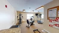 Located at 1320 Southwestern Blvd in Buffalo, NY, this single-floor fitness center offers a well-equipped workout space designed to support a variety of exercise routines. The room features warm beige walls, neutral carpeted flooring, and a bright, clean atmosphere illuminated by wood-trimmed ceiling light fixtures that lend a warm tone to the space.

The main workout area is outfitted with multiple cardio machines, including treadmills and a recumbent stationary bike, along with a multi-function cable strength training station and a rowing machine. A large wall-mounted mirror expands the sense of space and allows users to monitor their form during exercise. A wall-mounted flat-screen television is positioned for easy viewing from the cardio equipment, and a stability ball rounds out the functional fitness offerings.

Natural light filters in through windows fitted with horizontal blinds, providing a pleasant and energizing environment. A seating area along one side of the room includes a tufted black bench and a small table, offering a spot to rest or review workout notes. The space connects to an adjacent hallway through a white paneled door, suggesting convenient access to surrounding amenities. Overall, the fitness center provides a comfortable and practical environment suited for daily exercise.