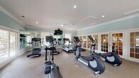 Located at 320 Glendare Dr in Winston-Salem, NC, this fitness center offers a spacious, single-level layout designed to accommodate a comprehensive workout experience. The space is finished with soft sage-green walls, crown molding, and a neutral-toned floor, creating a clean and polished aesthetic throughout.

The cardio area is generously outfitted with multiple treadmills, elliptical trainers, and additional aerobic machines arranged to allow ample space between equipment. A wall-mounted flat-screen television provides entertainment during workouts, while a ceiling fan contributes to airflow and comfort. Full-length mirrors along one wall visually expand the space and allow users to monitor their form.

The strength training section includes a cable machine, a weight bench, and a rack of color-coded kettlebells and free weights, offering versatile options for resistance training. Yoga mats are also available nearby.

Natural light fills the room through multiple windows and glass-paned French doors, which open to an outdoor area with views of a pool and surrounding greenery. Recessed lighting and ceiling-mounted fixtures ensure consistent illumination throughout. The French doors along one wall provide a seamless connection to an adjacent lounge or common area, enhancing the overall sense of openness and accessibility within the facility.