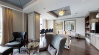 This single-floor space showcases a thoughtfully designed interior that blends classic architectural elements with contemporary elegance. The open layout connects a stylish lounge area with a separate dining or bar section, creating a cohesive and versatile environment.

The lounge area is anchored by a striking coffered ceiling detail with a recessed tray and a warm drum pendant light, adding depth and visual interest overhead. Dark hardwood flooring runs throughout, providing a rich contrast to the light, neutral wall tones. Seating includes a curved gray wingback chair with nailhead trim, a tufted cream sofa, and additional accent chairs, all arranged to encourage conversation and relaxation. A sculptural side table with a mirrored glass top adds a decorative focal point.

Adjacent to the lounge, a bar or dining area features tall upholstered counter chairs and pendant lighting, separated by a decorative column that maintains an open feel while subtly defining each zone. Large windows allow natural light to filter through sheer drapery. A framed botanical artwork and interior glass partition further enhance the refined, gallery-like ambiance of this well-appointed space.