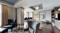This single-floor space showcases a thoughtfully designed interior that blends classic architectural elements with contemporary elegance. The open layout connects a stylish lounge area with a separate dining or bar section, creating a cohesive and versatile environment.

The lounge area is anchored by a striking coffered ceiling detail with a recessed tray and a warm drum pendant light, adding depth and visual interest overhead. Dark hardwood flooring runs throughout, providing a rich contrast to the light, neutral wall tones. Seating includes a curved gray wingback chair with nailhead trim, a tufted cream sofa, and additional accent chairs, all arranged to encourage conversation and relaxation. A sculptural side table with a mirrored glass top adds a decorative focal point.

Adjacent to the lounge, a bar or dining area features tall upholstered counter chairs and pendant lighting, separated by a decorative column that maintains an open feel while subtly defining each zone. Large windows allow natural light to filter through sheer drapery. A framed botanical artwork and interior glass partition further enhance the refined, gallery-like ambiance of this well-appointed space.