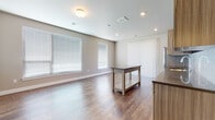 This single-floor residence offers a clean, modern aesthetic with a thoughtful layout designed for comfortable everyday living. The open-concept living and dining area is anchored by rich hardwood-style flooring and bathed in natural light from multiple large windows dressed with horizontal blinds. A sliding glass door with vertical blinds provides additional light and outdoor access.

The kitchen is a standout feature, showcasing contemporary flat-panel cabinetry in a warm wood-grain finish, stainless steel appliances including a side-by-side refrigerator with ice and water dispenser and a built-in dishwasher, a white subway tile backsplash, and an undermount sink. Open shelving adds a modern, functional touch. A kitchen island provides additional prep and dining space, flowing seamlessly into the main living area.

The residence includes two well-sized bedrooms, each finished with plush wall-to-wall carpeting and natural light from windows with blinds. Two bathrooms serve the home: one features a sleek framed glass walk-in shower with wood-look tile flooring, while the second offers a full soaking tub and shower combination. Both bathrooms include modern vanities with wood-grain cabinetry. Neutral warm-toned walls throughout the home contribute to a cohesive, inviting ambiance.