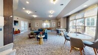 Located at 1045 Floribunda Way in Webster, NY, this single-floor community clubroom blends mid-century modern aesthetics with a warm, inviting ambiance. The space is thoughtfully designed to serve multiple social functions, flowing seamlessly from one area to the next across patterned carpeted floors.

The lounge area serves as a central focal point, anchored by a dramatic floor-to-ceiling geometric tile fireplace with a glossy metallic finish. A wall-mounted flat-screen TV, teal velvet seating, and a mid-century credenza complete this comfortable gathering spot. Adjacent to the lounge, a generously sized game area features a full-size pool table and a mounted Scrabble board, providing ample entertainment options.

The dining area is bathed in natural light through large windows dressed with floor-length drapes, offering views of the surrounding greenery. A round white dining table with upholstered chairs creates an intimate setting. Nearby, a sleek kitchenette features white cabinetry, a tiled teal accent wall, open shelving, and a white breakfast bar with wooden barstools — ideal for casual gatherings. Colorful mid-century artwork throughout ties the design together, creating a cohesive, vibrant, and welcoming community space.