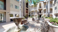 Located on Hollywood Boulevard in the heart of Los Angeles, this inviting outdoor courtyard showcases classic Mediterranean-inspired architecture with warm stucco facades, arched doorways, and wrought iron balconies spanning multiple levels above. The open-air courtyard serves as a central gathering space, anchored by ornate stone fountains that provide a serene, flowing water feature as a visual and auditory focal point.

Tall, slender palm trees rise dramatically through the courtyard, complemented by well-maintained raised garden beds brimming with colorful flowering plants, ferns, and tropical foliage. The hardscape features tasteful stone tile paving that extends throughout the courtyard, creating a cohesive and polished appearance.

Seating areas are thoughtfully arranged throughout the space, featuring classic wrought iron benches and bistro-style tables and chairs, ideal for relaxing or socializing outdoors. Decorative lantern-style sconces adorn the building walls, adding ambient lighting and architectural charm. Unit entryways are distinguished by arched tile accents and potted shrubs, enhancing the building's Mediterranean character. Abundant Southern California sunshine floods the courtyard, creating a bright, open atmosphere that highlights the lush greenery and architectural details throughout this well-appointed communal space.