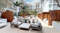 This elegant outdoor courtyard is a thoughtfully designed communal space enclosed by a modern, multi-story white building with expansive balconies on all sides. The space is defined by its resort-style atmosphere, combining refined materials and lush tropical landscaping to create a serene open-air retreat.

The courtyard is finished with a herringbone-patterned brick paver floor throughout, providing a warm and cohesive foundation. Generous seating areas are arranged across the space, featuring brushed metal-framed lounge chairs and sofas with plush white cushions, complemented by low concrete coffee tables. Warm wood slatted privacy screens and partitions divide the space into distinct zones, offering intimate pockets for gathering or relaxation.

Tall potted olive trees and palm trees are placed throughout, adding natural greenery and a Mediterranean character. A tiered garden feature anchors one end of the courtyard, incorporating ornate patterned tile risers, integrated raised planters filled with drought-tolerant succulents and tropical palms, and a sleek linear built-in fireplace as a focal point. Classic lantern-style outdoor light fixtures are positioned throughout, adding an elegant touch.

The fully open-air design allows abundant natural light to fill the space, while the surrounding building provides shelter and a sense of enclosure, making this courtyard an inviting and sophisticated outdoor living environment.