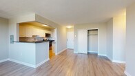 This single-floor unit is designed with a functional, open layout and finished consistently throughout with light hardwood-style flooring that adds warmth and continuity across all rooms. The living area is generously proportioned, featuring a large window fitted with blinds that frames outdoor views of surrounding greenery and neighboring buildings, allowing ample natural light to fill the space. A baseboard heating unit sits beneath the window, ensuring year-round comfort.

The kitchen is positioned in a semi-open configuration, accessible via a pass-through peninsula with dark countertops that contrasts cleanly against white cabinetry. The kitchen is fully equipped with stainless steel appliances, including a range, microwave, and dishwasher, along with abundant upper and lower cabinet storage and a stainless steel sink.

The bedroom features multiple windows offering outdoor views, a ceiling fan with integrated lighting, a dedicated closet with built-in shelving, and a baseboard heater. A second bedroom or additional room is also present, similarly outfitted with a ceiling fan and a reach-in closet.

The bathroom is finished with white cabinetry, a wall-mounted storage unit, wood-style flooring, and clean white wall paneling. The overall palette of neutral tones, white trim, and consistent flooring gives the space a cohesive and move-in-ready appearance.