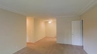 This single-floor residence offers a clean and classic interior defined by thoughtful architectural details and a cohesive design aesthetic. The living area is finished with wall-to-wall carpeting in a warm beige tone, creating a comfortable and inviting atmosphere underfoot. Crown molding runs along the ceiling perimeter, adding a refined touch to the otherwise understated space.

The walls are painted in a soft, neutral cream tone that enhances the sense of openness and pairs well with the natural and artificial lighting throughout. A flush-mount ceiling light fixture provides ambient illumination in the adjoining room, which opens seamlessly from the main living area, suggesting a connected, open-concept layout well-suited for both everyday living and entertaining.

A paneled interior door on one side of the space leads to an additional room, indicating a functional multi-room floor plan. Electrical outlets and light switches are conveniently positioned throughout, reflecting practical, well-considered design. The overall ambiance is calm and move-in ready, with neutral finishes that offer a versatile backdrop adaptable to a wide range of furnishing styles and personal tastes.
