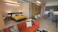 This single-floor space showcases a thoughtfully designed open-concept layout that seamlessly integrates a living area, sleeping zone, and dining space. Light wood-toned flooring runs throughout, lending warmth and visual continuity across the entire unit. A standout architectural feature is the full-height sliding barn door crafted from rich, natural wood paneling, which serves as a flexible divider between the sleeping and living areas, adding both functionality and a striking design element.

The sleeping area is anchored by a low-profile platform bed dressed in bold, colorful linens, complemented by a ceiling fan with ambient lighting overhead. The living area features a vibrant orange sofa with decorative throw pillows, creating an inviting focal point. Adjacent to the barn door, a compact dining nook accommodates a small round table with contemporary white chairs, ideal for casual meals. A potted plant introduces a touch of greenery, softening the modern aesthetic.

Flush-mounted lighting fixtures illuminate the space efficiently, while crisp white walls enhance the bright, airy atmosphere. A glimpse of a kitchenette can be seen toward the rear, completing this well-appointed, multifunctional layout.