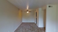 This single-floor Courtyard space features a open main living area finished with wall-to-wall carpeting in a neutral tone, providing a comfortable and practical foundation for furnishing. The room is topped with a textured ceiling and equipped with a ceiling fan fitted with a built-in light fixture, offering both air circulation and ambient lighting. The walls are painted in a soft, neutral hue that contributes to a calm and understated atmosphere throughout the space. A doorway visible toward the far end of the room leads to an additional area, suggesting a functional layout with clear separation between spaces. Architectural details include white trim and baseboards that provide clean contrast against the carpeted flooring and neutral walls. A visible HVAC vent in the ceiling indicates climate control is built into the unit. The overall design is straightforward and practical, with an emphasis on usability. The open feel of the main area allows for versatile furniture arrangements, making it adaptable to a variety of uses. The space presents a clean, move-in-ready environment suited for everyday living.