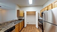 This single-story residence is designed with an open, functional layout that connects the living and kitchen areas seamlessly. The living room features warm wood-look flooring and a ceiling fan, creating a comfortable and well-ventilated space. Large vertical blinds along one wall allow for adjustable natural light, while an arched doorway adds an architectural detail that leads to the private areas of the home.

The kitchen is a standout feature, anchored by a peninsula with a dark granite-style countertop that provides both prep space and a casual dining or serving surface. Honey-toned wood cabinetry lines the walls, offering generous storage, while stainless steel appliances — including a refrigerator, gas range, and microwave — add a modern touch. A double-basin sink and a dedicated laundry closet with bifold doors are also integrated into the kitchen. Wood-look flooring continues throughout this space for easy maintenance.

The bedroom is carpeted and spacious, with a ceiling fan and a window that allows natural light and outdoor views. The bathroom features warm wood cabinetry, a single-sink vanity with a wide mirror, and a full tub-shower combination with white tile surround.