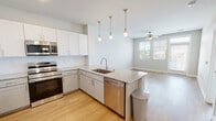 This well-appointed single-floor residence showcases a bright, open-concept layout that seamlessly connects the kitchen and living areas. The kitchen is a standout feature, boasting crisp white shaker cabinetry, light granite countertops, and a full suite of stainless steel appliances, including a side-by-side refrigerator with water dispenser, electric range, built-in microwave, and dishwasher. A peninsula island with an undermount sink serves as the centerpiece, illuminated by a trio of elegant gold-toned pendant lights that add a warm, contemporary touch. Light hardwood flooring flows throughout the kitchen and living space, enhancing the sense of openness and brightness. The adjacent living area benefits from natural light streaming through a large window fitted with blinds, complemented by a ceiling fan overhead. The bedroom is a comfortable retreat, featuring plush carpeting, a ceiling fan, and a window that brings in natural light. The bathroom maintains the home's consistent design language with a granite-topped vanity, a large mirror, and a glass-enclosed walk-in shower, all finished in clean, neutral tones. Throughout the space, light-toned walls and natural light create a fresh, airy ambiance.