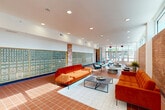 Located at 8 S Sixth St in Council Bluffs, Iowa, this single-floor lobby and communal living space offers a thoughtfully designed environment that blends mid-century character with modern functionality. The expansive open floor plan is anchored by warm terracotta square tile flooring that extends throughout the entire space, complemented by crisp white area rugs that define the seating areas.

A defining architectural feature is the full-length exposed brick wall running along one side, adding texture and warmth to the interior. Large floor-to-ceiling glass entry doors and a continuous band of clerestory windows flood the space with abundant natural light, offering views of the surrounding outdoor greenery. Recessed ceiling lighting supplements the natural light throughout.

A prominent wall of individual mailboxes lines one side of the room, a hallmark feature indicating this serves as a central gathering and reception hub for a multi-unit building. Brick columns punctuate the space, reinforcing the structural character of the design.

The living area is furnished with bold orange velvet sofas, teal accent chairs, and low-profile modern coffee tables, creating a vibrant yet functional seating arrangement. Additional seating clusters toward the entrance provide flexible gathering opportunities. The overall ambiance is welcoming, bright, and community-oriented, making it an inviting space for residents and visitors alike.