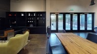 This single-floor communal space blends contemporary design with an artistic, urban aesthetic to create a dynamic and inviting environment. The open-plan layout is anchored by rich dark hardwood flooring throughout, complemented by high ceilings that lend an expansive, airy feel to the interior.

Upon entry, a glass-paneled door floods the foyer with natural light, where a wall-mounted bank of flat-screen televisions sits above a sleek, dark credenza — ideal for media viewing or presentations. Bold, large-scale mixed-media artwork adorns the walls, infusing the space with energy and creative character.

The main gathering area features multiple distinct seating zones. A pair of cognac leather barrel chairs rest atop a striped area rug near floor-to-ceiling glass doors, offering views of an outdoor terrace. A vibrant teal sectional sofa provides a lively contrast against the neutral palette, positioned adjacent to a communal wood-topped table with mixed seating including ghost chairs and upholstered dining chairs.

A striking black built-in shelving unit lines one wall, displaying curated decorative objects and serving as a functional backdrop. Pendant globe lighting and track lighting overhead provide warm, layered illumination throughout the space. Multiple sets of full-glass exterior doors connect the interior to the outdoors, ensuring the space remains bright and well-connected to its surroundings.