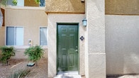 This single-floor unit is accessed through a distinctive green six-panel door set within a textured stucco exterior, complemented by a wall-mounted lantern light fixture. The exterior's two-tone stucco finish and desert landscaping establish a southwestern architectural character consistent with the surrounding community.

Inside, the open-concept layout connects the living area and kitchen in a seamless, airy arrangement. The kitchen features warm honey-toned wood cabinetry extending to upper cabinet heights, offering generous storage. A peninsula with a light-colored countertop serves as both a workspace and casual dining counter, separating the kitchen from the living area. Appliances include a white refrigerator, microwave, and dishwasher. Overhead, a track lighting fixture illuminates the space efficiently.

The living area is finished with neutral beige carpeting and light gray walls, creating a clean, understated palette throughout. A window on the far wall draws in natural light, brightening the open floor plan. Ceiling-mounted HVAC vents ensure comfortable climate control.

A private balcony extends the living space outdoors, offering views of mature palm trees, landscaped grounds, and the surrounding community — a notable highlight that brings a resort-like quality to everyday living.