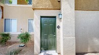 This single-floor unit is accessed through a distinctive green six-panel door set within a textured stucco exterior, complemented by a wall-mounted lantern light fixture. The exterior's two-tone stucco finish and desert landscaping establish a southwestern architectural character consistent with the surrounding community.

Inside, the open-concept layout connects the living area and kitchen in a seamless, airy arrangement. The kitchen features warm honey-toned wood cabinetry extending to upper cabinet heights, offering generous storage. A peninsula with a light-colored countertop serves as both a workspace and casual dining counter, separating the kitchen from the living area. Appliances include a white refrigerator, microwave, and dishwasher. Overhead, a track lighting fixture illuminates the space efficiently.

The living area is finished with neutral beige carpeting and light gray walls, creating a clean, understated palette throughout. A window on the far wall draws in natural light, brightening the open floor plan. Ceiling-mounted HVAC vents ensure comfortable climate control.

A private balcony extends the living space outdoors, offering views of mature palm trees, landscaped grounds, and the surrounding community — a notable highlight that brings a resort-like quality to everyday living.