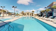 This expansive resort-style outdoor amenity area is designed to deliver a vacation-like experience. At its centerpiece is a large, crystal-clear swimming pool with a gradual entry and in-water sun shelves topped with loungers, perfect for relaxing while partially submerged. Adjacent to the main pool sits a bubbling spa, offering a more intimate retreat. The surrounding pool deck is generously appointed with rows of white chaise loungers, shaded umbrella stations, and teal-accented furnishings that lend the space a vibrant, cohesive aesthetic. Tall palm trees are strategically placed throughout, evoking a tropical atmosphere under open blue skies. A dedicated cabana area features semi-enclosed structures with translucent roofing, mesh screens, turf flooring, and striped lounge chairs — offering a private, shaded escape from the sun. The outdoor social zone includes a sweeping curved sectional sofa upholstered in bold teal, arranged around a central table on a stone-tiled patio, ideal for gatherings. An outdoor kitchen station, fitted with stainless steel grills, stone-clad countertops, and a pergola overhead, completes the entertaining amenities. The entire space is professionally landscaped and designed for both relaxation and social activity.