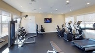 The fitness center at Beacon Place is a well-appointed, single-floor exercise facility designed to support a well-rounded workout routine. The room features a dark, durable flooring surface that provides a functional foundation for both cardio and strength training activities. Recessed lighting throughout the ceiling ensures consistent, even illumination across the entire space, complementing the natural light that filters in through windows along the walls.

The cardio area is equipped with multiple elliptical machines and a stationary bike, all positioned conveniently near the windows. A wall-mounted flat-screen television is centrally placed, offering entertainment during workouts. On the opposite side of the room, a dumbbell rack holds a range of free weights alongside a weight bench, catering to strength training needs. A cable machine adds versatility for a variety of resistance exercises.

The walls are finished in a neutral, warm tone that contributes to an open and inviting atmosphere. A white paneled door provides access, while the overall layout allows for comfortable movement between equipment. The space is clean, organized, and thoughtfully arranged to accommodate multiple users simultaneously.