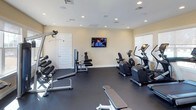 The fitness center at Beacon Place is a well-appointed, single-floor exercise facility designed to support a well-rounded workout routine. The room features a dark, durable flooring surface that provides a functional foundation for both cardio and strength training activities. Recessed lighting throughout the ceiling ensures consistent, even illumination across the entire space, complementing the natural light that filters in through windows along the walls.

The cardio area is equipped with multiple elliptical machines and a stationary bike, all positioned conveniently near the windows. A wall-mounted flat-screen television is centrally placed, offering entertainment during workouts. On the opposite side of the room, a dumbbell rack holds a range of free weights alongside a weight bench, catering to strength training needs. A cable machine adds versatility for a variety of resistance exercises.

The walls are finished in a neutral, warm tone that contributes to an open and inviting atmosphere. A white paneled door provides access, while the overall layout allows for comfortable movement between equipment. The space is clean, organized, and thoughtfully arranged to accommodate multiple users simultaneously.