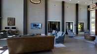 This impressive single-floor space commands immediate attention with its soaring ceilings and grand architectural scale. Tall, dark structural columns frame the expansive main living area, drawing the eye upward along smooth, neutral-toned wall panels that lend a clean, contemporary aesthetic. A statement chandelier with warm, ambient lighting anchors the upper space, adding a touch of classic elegance to the modern design.

The main gathering area is thoughtfully arranged with a rich leather sofa and a coordinated set of upholstered armchairs in soft blue-gray tones, creating an inviting and balanced seating arrangement. A flat-screen television is mounted flush against the feature wall, seamlessly integrated into the refined décor. Large windows and glass-panel doors allow natural light to filter through, connecting the interior to the outdoors and enhancing the open, airy atmosphere.

The neutral color palette throughout — from the light stone-toned flooring to the textured wall surfaces — creates a cohesive and calming environment. The generous proportions of the space make it well-suited for both relaxed everyday use and larger social gatherings. Overall, this space delivers a harmonious blend of architectural drama and comfortable, well-curated design.