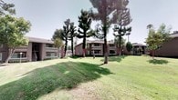 This ground-floor unit is part of a charming multi-building residential community defined by its expansive, manicured green spaces and mature landscaping. Towering pine and shade trees are distributed throughout the grounds, creating a park-like atmosphere that adds a sense of privacy and natural beauty to the community. Well-kept lawns stretch between the buildings, complemented by neatly trimmed hedges, ivy ground cover, and brick-edged garden beds that line the walkways. Smooth concrete paths wind through the community, connecting the buildings and leading to a shared pool area, which features patio seating shaded by umbrellas and is enclosed by a classic white fence. The residential buildings themselves are constructed with a combination of stucco exteriors, brick accents, and wood trim details, lending a warm and cohesive architectural character. Exterior staircases with painted railings provide access to upper levels, while ground-floor units benefit from covered entryways and direct access to the surrounding grounds. Safety features such as wall-mounted lighting and accessible fire extinguisher stations are thoughtfully integrated throughout. The overall ambiance is one of a serene, community-oriented environment with ample outdoor space and thoughtful maintenance.