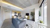This single-floor space showcases a thoughtfully designed open-concept layout that integrates the living area and kitchen into one cohesive environment. The exposed concrete ceiling serves as a defining architectural feature, lending an industrial character balanced by warm ambient lighting from a recessed cove that runs along the ceiling's perimeter. Polished concrete floors extend throughout, reinforcing the contemporary aesthetic while providing a durable, low-maintenance surface.

The kitchen is outfitted with light wood cabinetry, stainless steel appliances including a refrigerator, microwave, and range, and a breakfast bar with seating for two. The clean white countertops provide a bright contrast against the warm cabinetry tones.

The living area accommodates a comfortable gray sectional sofa, anchored by a patterned area rug. Decorative accents, including a globe-style lamp and a vibrant yellow accent cabinet, introduce personality and color to the otherwise neutral palette. Lush potted plants add a natural, organic element to the space. The open layout and cohesive design create a functional, modern living environment with an urban, sophisticated ambiance.