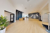 Located at 124 Maple St in Danvers, MA, this expansive lobby presents a refined, contemporary design across a generously sized single floor. The entrance immediately impresses with light hardwood flooring that extends throughout, complemented by soaring white ceilings with recessed lighting that enhance the sense of openness and airiness.

At the heart of the space sits a sculptural reception desk framed by a dramatic navy blue slatted feature wall with integrated LED strip lighting, creating a bold architectural focal point. Opposite, a slate-gray accent wall adorned with vertical gold pendant rods adds warmth and visual rhythm, anchoring a sophisticated lounge seating area complete with a flat-screen TV, plush sofas, and textured area rugs.

A dedicated seating alcove features a marble-tiled feature wall, tufted banquette seating, natural wood stump side tables, and floor-to-ceiling sheer-draped windows that flood the space with natural light and offer views of the surrounding streetscape. An open-cage bookshelf display unit and additional lounge groupings with warm-toned furnishings enrich the communal areas.

The hallway connects seamlessly to elevators and adjacent rooms, with luggage carts and clean sightlines reinforcing the space's functional, hospitality-forward character. Overall, the lobby balances striking design details with a welcoming, well-lit atmosphere.
