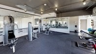 This fitness center offers a comprehensive workout environment designed to support a wide range of exercise routines. The space features an industrial-inspired aesthetic, with exposed concrete ceilings, visible ductwork, and track lighting that lend a modern, utilitarian character throughout.

The main workout floor is finished with durable dark carpet tile and is generously sized to accommodate multiple exercise stations simultaneously. A dedicated cardio zone is equipped with a treadmill, elliptical trainer, and a spin bike, all positioned along a windowed wall that allows natural light to pour in and provides views to the outdoors. A ceiling fan and wall-mounted air conditioning unit ensure comfortable ventilation during workouts.

The strength training area features a selection of cable and resistance machines, along with additional selectorized equipment, all reflected in a large wall-mounted mirror that visually expands the space. A stair climber rounds out the cardio offerings.

A separate functional training room provides a bright, open area with speckled flooring, a rack stocked with medicine balls of varying weights, resistance bands, and a stability ball, making it ideal for dynamic and rehabilitative exercise. A storage room is also accessible from this area. Natural light from large windows enhances the open feel of this zone, creating an inviting and energizing atmosphere throughout the facility.