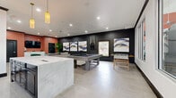 This single-floor amenity space is thoughtfully designed to blend social functionality with contemporary style. The open layout flows seamlessly between distinct zones, each characterized by bold design choices and high-quality finishes throughout.

The game area serves as a central focal point, anchored by a full-size pool table set against a dramatic dark accent wall adorned with a curated series of large abstract art pieces in navy, gold, and blush tones. A foosball table adds to the recreational offerings, while recessed lighting keeps the space well-lit and inviting.

Adjacent to the game area, a wet bar and kitchenette provides excellent hosting capabilities. Dark cabinetry with brushed gold hardware lines the walls beneath a marble-style countertop, complemented by a patterned tile backsplash in soft sage tones. A built-in beverage refrigerator and a large central island with seating complete this well-appointed prep area. Elegant fringe pendant lights in amber glass add a warm, artistic touch overhead.

The lounge area features plush seating including a charcoal sofa, emerald green velvet accent chairs, and a marble-top coffee table. A wall-mounted television, terracotta accent walls, and lush tropical plants create a warm, vibrant atmosphere. Large format neutral floor tiles run throughout, tying each zone together into a cohesive, polished aesthetic.