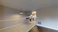 This single-floor residence offers a practical and comfortable layout designed for everyday living. The open-concept design connects the living area and kitchen, creating a cohesive and functional space. The kitchen is equipped with warm wood-toned cabinetry, a dark countertop peninsula that doubles as a breakfast bar, a white refrigerator, stove, and built-in microwave. A flush ceiling light illuminates the kitchen, while a stylish industrial-style pendant lamp hangs over the transition into the living area, adding a touch of character. The flooring transitions from light beige ceramic tile in the kitchen and dining area to rich hardwood in the living space, clearly delineating each zone. The walls are painted in a neutral gray tone, lending the interior a clean and modern feel. A floor vent is visible, indicating a central HVAC system for year-round climate control. The overall ambiance is understated and practical, with a straightforward layout that maximizes usability. The open sightlines between the kitchen and living area make the space feel airy and well-connected, well-suited for both daily routines and casual entertaining.
