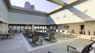 This rooftop terrace offers a sophisticated open-air retreat elevated above the surrounding cityscape. At the heart of the space is a sleek, granite-clad water feature with cascading fountain jets, creating a calming focal point surrounded by a shallow reflection pool. The deck is finished in composite wood-tone planking, lending a warm, contemporary feel underfoot.

Ample lounge seating is arranged throughout the terrace, with sling-style chaise lounges and chairs positioned to take advantage of both the water feature and the panoramic urban views. Multiple seating clusters provide flexible arrangements for relaxation or social gatherings. Overhead, a series of clean architectural pergola beams frame the open sky, adding structure and visual interest without obstructing the sweeping views of the city skyline beyond.

The surrounding building façade features a modern neutral palette of grey and beige paneling, contributing to the terrace's polished, urban aesthetic. Additional amenity areas line the perimeter, including what appears to be a bar or service counter and supplementary lounge groupings. The space benefits from abundant natural light throughout the day, making it a dynamic environment suited for both daytime relaxation and evening gatherings.
