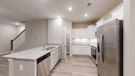 This two-story residence is designed with a clean, contemporary aesthetic throughout. The kitchen serves as a functional centerpiece on the main level, showcasing white shaker-style cabinetry with crown molding detail, light granite countertops, and a full suite of stainless steel appliances including a side-by-side refrigerator, electric range, over-the-range microwave, and dishwasher. Light wood-look tile flooring runs through the kitchen, complementing the neutral warm-gray wall tones and recessed lighting overhead. A convenient half bath on the main level features wood-look flooring and a pedestal sink. Upstairs, the primary bedroom is notably spacious, featuring plush carpeting, a tray ceiling with crown molding, and a ceiling fan. Additional bedrooms are carpeted and receive natural light through double-hung windows with blinds. A full bathroom upstairs offers marble-look tile flooring, a tub-shower combination, white cabinetry, and brushed nickel fixtures. A dedicated laundry room is equipped with wire shelving, washer/dryer hookups, and a utility connection panel, adding practical convenience to the upper level.