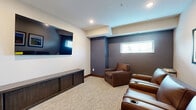 Located at 1415 County Road B W in Roseville, MN, this dedicated video room is a compact, thoughtfully designed space built for comfortable media viewing. The room features a neutral-toned, textured carpet that adds warmth underfoot, while the walls are finished in a combination of crisp white and deep slate blue, creating a cozy, immersive atmosphere well-suited for screening sessions. A large wall-mounted flat-screen television serves as the focal point of the room, positioned above a low-profile dark wood media console with cabinet storage. Seating consists of multiple brown leather recliners and armchairs equipped with cup holders, arranged to face the screen for an optimal viewing experience. Recessed ceiling lighting provides ambient illumination throughout the space. A horizontal window fitted with blinds along one wall allows for controlled natural light. Framed photographic artwork depicting city and waterfront scenes — reflecting the local Twin Cities area — adorns the wall alongside the seating area, adding a regional character to the room. A frosted-glass interior door with a numbered room placard provides a semi-private entry. The overall ambiance is intimate and purposefully functional, making this video room a well-appointed retreat for entertainment and relaxation.
