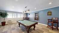 Located in Escondido, CA, this dedicated billiards room is a purpose-built game space designed for recreational enjoyment. The room is centered around a full-size pool table with a green felt surface and rich mahogany-finished legs, positioned beneath a striking three-shade pendant light fixture that provides focused, even illumination over the playing surface. Crown molding runs along the perimeter of the ceiling, lending a polished, finished look to the space.

The walls feature a two-tone design, with a calming blue accent wall providing a visual backdrop to the pool table and its accessories. A wall-mounted cue rack keeps billiards equipment neatly organized, while decorative pieces including a clock and game-themed wall art add character to the room. A high-top table with bar stools offers comfortable seating for those waiting their turn or simply observing play.

Patterned carpeting in neutral earth tones covers the floor, contributing to the room's casual yet well-maintained atmosphere. Windows along one wall allow natural light to filter in, brightening the space during daytime hours. Decorative potted trees placed near the arched entryway add a touch of greenery and warmth. A glass-paneled door leads to an adjoining area, making this game room a well-connected and inviting recreational retreat.