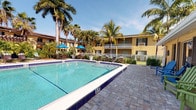 This inviting outdoor amenity area showcases a generously sized swimming pool accented by classic blue tile trim, offering both shallow and deeper sections accessible via stainless steel entry rails. The pool is surrounded by a well-maintained paver stone deck that provides ample space for lounging and socializing. Colorful Adirondack chairs in blue and green are arranged along the poolside, adding a cheerful, resort-style atmosphere to the setting.

The space is framed by a white metal security fence and lush tropical landscaping, including tall swaying palm trees that lend a distinctly Florida ambiance. Blue patio umbrellas with accompanying seating provide shaded areas ideal for relaxing on sunny days. A hot tub is conveniently situated at one end of the pool area, while a dedicated outdoor grilling and BBQ station adds practical appeal for gatherings.

Adjacent to the pool, a single-story stucco building with black shutters and French doors serves as a pool house or community facility, with a life ring mounted on the exterior wall for safety. The warm yellow exterior walls of the surrounding multi-story building complement the tropical setting. Overall, the space exudes a relaxed, resort-inspired character with thoughtful amenities designed for outdoor leisure and community enjoyment.