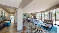 This well-appointed single-story community clubhouse offers a thoughtfully designed layout with multiple functional zones that seamlessly flow into one another. The main lounge area is bright and open, furnished with contemporary patterned chairs, a plush sofa accented with teal throw pillows, and a wooden coffee table, all anchored by a teal area rug. Floor-to-ceiling sliding glass doors line one wall, flooding the space with natural light and providing direct views of an outdoor patio area shaded by umbrella-covered seating. A billiards table serves as a centerpiece for recreational use, complemented by bar-height seating nearby and a wall-mounted television. The communal kitchen features stainless steel appliances including a side-by-side refrigerator, a mosaic tile backsplash, sleek wood-grain cabinetry, and a large white countertop island with bar stool seating—all beneath recessed lighting and wall-mounted screens. A dedicated business center provides multiple workstations along wall-mounted desks, ergonomic seating, and teal accent walls adorned with framed artwork. The entry corridor offers a welcoming transition space with built-in bench seating, round side tables, wall sconces, and large-scale photographic wall art, creating an inviting passage that connects the various areas of this versatile community facility.