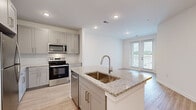 Located at 550 Webb Gin House Rd in Lawrenceville, GA, this single-floor residence offers a thoughtfully designed open-concept layout with contemporary finishes throughout. The kitchen is a standout feature, showcasing granite countertop surfaces, a central island with an undermount sink and pull-down faucet, soft gray shaker-style cabinetry with brushed gold hardware, a subway tile backsplash, and a full suite of stainless steel appliances including a refrigerator, range, over-the-range microwave, and dishwasher. Light wood-style flooring runs through the main living areas, complementing the bright, neutral color palette. The living area flows naturally from the kitchen and opens to a private balcony through glass-paned doors, offering views of the surrounding community and tree line beyond. Bedrooms are carpeted for comfort and feature natural light through window treatments. A walk-in closet with wire shelving provides generous storage. The bathroom is finished with granite countertops, gray cabinetry, a large mirror, and a tub-shower combination, all set on light wood-style flooring. Recessed lighting throughout the unit adds a clean, modern ambiance to every room.