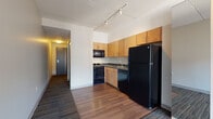 This single-floor unit presents a practical, modern layout with distinct functional zones flowing from the entryway through to the living space. The kitchen is positioned at the front of the unit and is equipped with warm-toned wood cabinetry extending to the ceiling for generous storage. Black appliances—including a gas range, over-the-range microwave, dishwasher, and full-size refrigerator—contrast neatly against dark countertops and hardwood-style flooring. Track lighting overhead provides directed illumination throughout the cooking area. The open layout transitions into a carpeted living area featuring multi-tone loop-pile carpet, creating a visual boundary between the kitchen and the main living zone. A large window at the far end of the living space brings in natural light and offers views of the surrounding urban environment. A hallway leads to the bathroom, which features light tile flooring, a wood-cabinet vanity with a white countertop and wall mirror, grab bars, and a separate walk-in shower stall with a full-height partition door. The overall design is clean and understated, characterized by white walls, practical finishes, and a straightforward, functional aesthetic suited for everyday living.