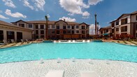Located at Avalon Point in Missouri City, Texas, this single-story amenity space is designed to deliver a resort-like experience within a well-appointed residential community. The centerpiece of the outdoor area is a large, shimmering pool with crystal-clear water, accented by fountain jets and a shallow entry zone that creates an inviting, vacation-style atmosphere. Surrounding the pool, a generously sized paver deck provides ample room for lounge chairs arranged across both turf and stone surfaces, offering versatile spaces for sunbathing and relaxation. Towering palm trees are strategically placed throughout the grounds, reinforcing the tropical aesthetic. A pergola-shaded seating area and a fire pit zone further extend outdoor entertaining possibilities.

Adjacent to the pool deck, a covered lounge area features smooth tile flooring, recessed lighting, and floor-to-ceiling glass walls and sliding doors that frame unobstructed views of the pool and surrounding buildings. This transitional indoor-outdoor space is furnished with dark wicker sectionals and chairs accented with vibrant teal cushions, creating a modern, welcoming ambiance. The stucco exterior and clean architectural lines of the community buildings complete the refined, upscale character of this well-designed communal space.