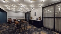 Located at 1333 N Santa Fe Ave in Vista, California, this single-floor space offers a thoughtfully designed interior that blends modern industrial aesthetics with warm, inviting elements. The main area is anchored by a striking ceiling grid of brass-finished track lighting adorned with exposed Edison-style bulbs, casting a warm, ambient glow throughout. Glossy white subway tile backsplash adds a clean contrast against the dark, sleek cabinetry topped with a white countertop, which serves as a functional credenza area complete with drawers and decorative greenery. A large projection screen mounted on the wall makes this space well-suited for presentations or screenings, complemented by a high-top communal table surrounded by industrial-style bar stools. The patterned carpet—featuring dramatic charcoal, gold, and cream tones—anchors the space with visual depth and texture. A recessed lounge nook with a dark ceiling, teal-blue walls, and geometric patterned wallpaper offers a more intimate setting, furnished with colorful woven rocking chairs, a live-edge wood table, and a plush banquette. Floor-to-ceiling glass partition walls with black framing allow visibility between areas while maintaining a sense of defined zones. The overall ambiance is contemporary and versatile, ideal for collaborative gatherings, private events, or creative meetings.