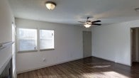 This single-floor residence offers a clean, open layout designed for comfortable everyday living. The main living area is a standout feature, showcasing rich, dark hardwood-style flooring that runs throughout and provides a warm contrast against the crisp white walls. A textured ceiling adds subtle architectural character to the space, while a ceiling fan with an integrated light fixture ensures year-round comfort and practical illumination. Two windows fitted with horizontal blinds line one wall, allowing natural light to fill the room and create a bright, welcoming atmosphere. A flush-mount ceiling light near the entry provides additional overhead lighting. The entry door, finished in a soft neutral tone, opens directly into the living area, creating a seamless transition from the outside in. The space presents a neutral, move-in-ready palette throughout, offering a versatile backdrop that accommodates a wide range of interior styles and furnishings. The open, uncluttered floor plan maximizes the sense of space, making the living area feel expansive and airy. Overall, this residence combines practical design elements with a clean aesthetic to deliver a functional and inviting living environment.
