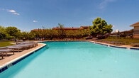 Located at 201 Idlewild Rd in Bel Air, Maryland, this outdoor pool facility serves as a central community amenity within a well-maintained residential complex. The pool itself is expansive, featuring clear turquoise water, dedicated lap lanes, and multiple entry points via stainless steel ladders. The pool deck is generously sized, offering ample space for circulation and relaxation around the perimeter.

The sun deck is outfitted with rows of modern sling-style chaise lounges arranged neatly along the poolside, providing residents with comfortable spaces to unwind and enjoy the open sky. A black iron perimeter fence encloses the entire pool area, ensuring both safety and a defined boundary between the pool grounds and the surrounding green space.

Well-maintained lawns extend beyond the fencing, lending a park-like quality to the environment. Mature trees provide natural shade and a pleasant visual backdrop throughout the space. A dedicated pool house structure with a distinctive cupola roofline is situated adjacent to the pool, likely serving as a support facility. The surrounding multi-story residential buildings are visible in the background, contextualizing the pool as a shared community resource. Under clear blue skies, the entire area projects a bright, welcoming, and resort-inspired atmosphere ideal for warm-weather recreation.