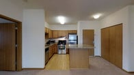 This single-floor residence offers a thoughtfully designed open-concept layout that seamlessly connects the kitchen and living areas. The kitchen is a functional centerpiece, outfitted with warm oak cabinetry that provides generous storage, granite-patterned countertops, and a full suite of stainless steel appliances including a gas range, refrigerator, and dishwasher. A built-in microwave adds everyday convenience. A peninsula island with matching countertop extends the workspace and serves as a natural divider between the kitchen and the adjacent living area, making it ideal for casual dining or entertaining. The kitchen features easy-care light hardwood flooring, while the living space transitions to plush carpeting, adding warmth and comfort underfoot. Flush-mount ceiling lighting provides consistent, even illumination throughout. The entry door, finished in a coordinating wood tone, contributes to the cohesive and inviting aesthetic of the space. Overall, the residence presents a clean, practical, and comfortable environment well-suited for everyday living.