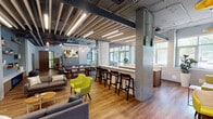 Located at 3233 S Dr Martin Luther King Jr Dr in Chicago's Bronzeville neighborhood, this single-floor space showcases a thoughtfully designed industrial-chic interior that balances open communal areas with functional amenities. The architecture retains raw, expressive elements throughout, including exposed concrete columns, visible ductwork and piping along the ceilings, and structural steel accents, all of which contribute to a distinctly urban aesthetic.

The main lounge and social area features warm hardwood-style flooring, high ceilings, and abundant natural light streaming through large, multi-pane windows that frame views of the surrounding tree-lined streetscape. Seating is varied and plentiful, with vibrant chartreuse accent chairs and sofas, modular sectionals, and a large U-shaped lounge configuration anchored beneath a statement drum pendant light. Bold wall murals and colorful artwork add character throughout.

A central communal zone offers bar-height seating along a counter, a dining table with mixed seating, and a kitchen-style service area, making it well-suited for both casual gatherings and collaborative use. Glass-paneled partitions provide subtle separation between zones without sacrificing the open, airy feel.

The on-site fitness center is equipped with cardio machines including treadmills and ellipticals, free weights, TRX suspension systems, and mirrored walls, all set on durable rubberized flooring beneath industrial-style linear lighting.
