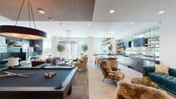 This single-floor amenity space is a masterfully designed social hub that seamlessly blends entertainment, relaxation, and refined aesthetics. The open-plan layout connects several distinct zones, each with its own character while maintaining a cohesive, upscale atmosphere throughout.

At the heart of the space sits a regulation billiards table, anchored beneath a striking candle-style chandelier. Adjacent to it, a dedicated music alcove houses a sleek black grand piano, accompanied by a tufted teal velvet sofa, framed black-and-white photography, and plush faux-fur lounge chairs — creating an intimate, gallery-like retreat.

The full wet bar serves as a central gathering point, featuring a dark countertop bar with upholstered seating, a brass overhead wine glass rack, open shelving, and mirrored cabinetry. An industrial-style linear chandelier adds warmth and visual drama overhead.

The lounge seating area is generously appointed with tan wingback chairs, a glass-and-brass coffee table, and potted olive trees that bring life and texture to the space. Floor-to-ceiling windows along multiple walls flood the interior with natural light and offer expansive urban skyline views.

A moody private dining nook with dark wood paneling, curved banquette seating in charcoal leather, a marble-top table, and a brass dome pendant light provides an intimate contrast to the open social areas. Light hardwood flooring ties the entire space together elegantly.
