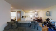 This single-floor residence offers a practical and comfortable open-concept layout that seamlessly connects the main living areas. The living room serves as the central gathering space, furnished with a large, plush sectional sofa that anchors the room. A ceiling fan with a light fixture provides both ventilation and ambient lighting overhead.

The kitchen flows directly from the living area and is equipped with white cabinetry offering generous storage, a white electric range with overhead ventilation, and a full-size refrigerator. The countertops provide ample workspace, and the layout is designed for efficient meal preparation.

Adjacent to the kitchen, a dedicated dining area accommodates a wooden table and matching chairs, making it a functional space for everyday meals. The dining and kitchen zones are unified by consistent flooring and an open sightline that enhances the sense of space throughout.

A doorway visible from the living area leads to a bedroom, which features carpeted flooring and a standard closet door, offering a private retreat from the shared living spaces. The overall ambiance of the home is warm and approachable, with neutral wall tones and practical finishes that create a welcoming, lived-in atmosphere throughout.
