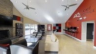 This single-floor communal space is designed with a contemporary aesthetic, highlighted by a dramatic vaulted ceiling that rises to a prominent peak, creating an airy and open atmosphere throughout. Two ceiling fans are mounted overhead, ensuring comfortable airflow across the expansive room.

The interior is divided into distinct functional zones. A comfortable lounge area anchors the center of the space, furnished with a cream leather sofa and dark upholstered seating arranged around a low coffee table, ideal for relaxation or socializing. Adjacent to this is a high-top table and chair seating area positioned near large sliding glass doors, which flood the space with natural light and provide direct access to an outdoor patio.

A dedicated computer workstation area lines the far wall, featuring multiple monitors on bar-height desks with stools, accented by a bold red accent wall with a geometric red, black, and gray panel display. A bookcase along one wall adds additional utility to the space.

The overall color palette blends neutral beige walls with striking red accents, polished light flooring, and recessed lighting, creating a vibrant yet functional environment suited for both work and leisure.
