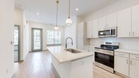 This thoughtfully designed one-bedroom, one-bathroom residence offers a clean, modern aesthetic across a single floor of comfortable living space. The open-concept kitchen serves as the heart of the home, anchored by a large center island with a white quartz countertop and an integrated sink with a pull-down faucet. Elegant gold-accented pendant lights hang above the island, complementing the white shaker cabinetry and herringbone tile backsplash. Stainless steel appliances, including a French door refrigerator, range, and built-in microwave, provide a fully equipped culinary space. Light wood-style flooring flows throughout the main living area, contributing to a bright and airy feel. Natural light pours in through large windows and a glass-panel entry door, offering pleasant outdoor views. The bedroom features plush carpeting, a generously sized window with tree-lined views, and access to a spacious walk-in closet outfitted with wire shelving systems for ample organization. The bathroom is finished with herringbone wall tile surrounding a full soaking tub and shower combination, paired with wood-look flooring for a cohesive, contemporary look throughout.